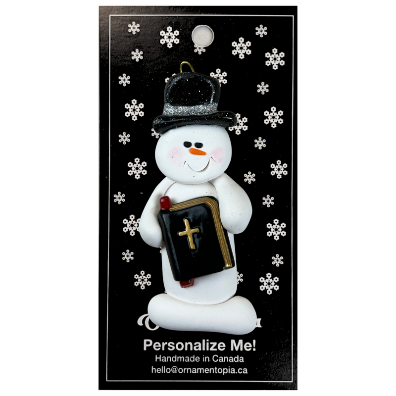 Ornament of white snowman in black top hat, holding a Bible, on display backing card.