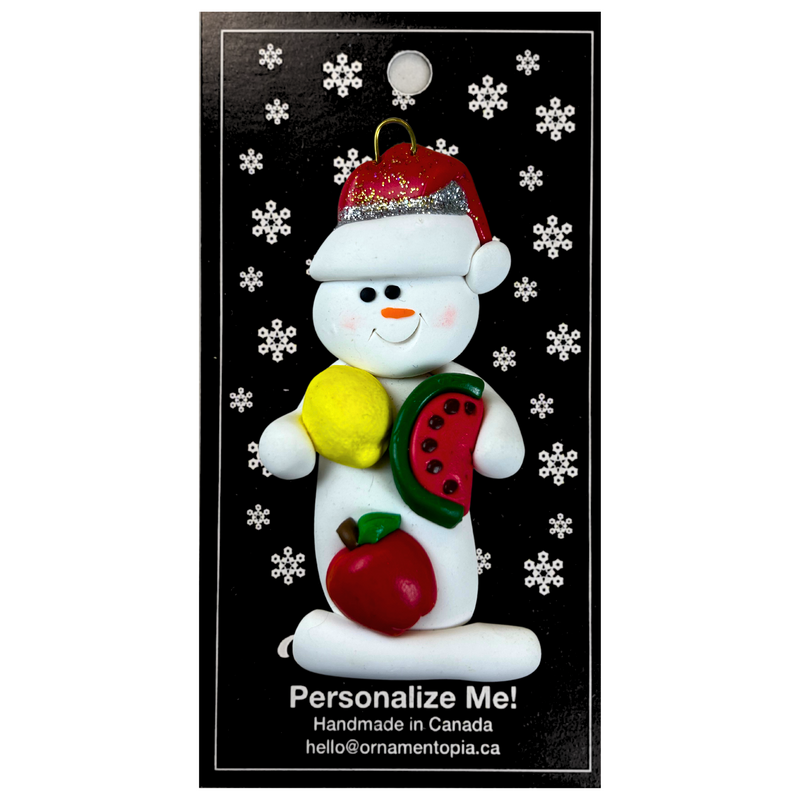 Ornament of white snowman in red hat holding various fruits, on display backing card.