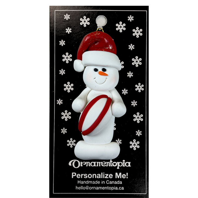 Ornament of white snowman in red hat holding a red and white rugby ball, on display backing card.