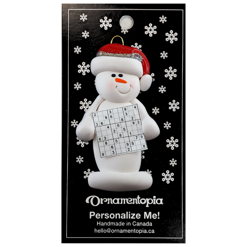 Ornament of white snowman in red hat holding a sudoku page, on display backing card.
