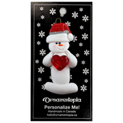 Ornament of white snowman in red hat holding a shiny red heart, on display backing card.