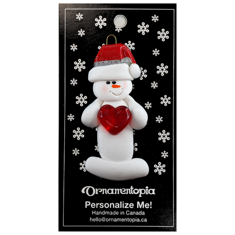 Ornament of white snowman in red hat holding a shiny red heart, on display backing card.