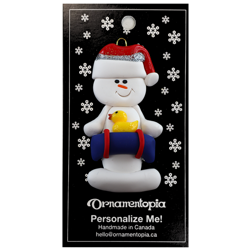 Ornament of white snowman in red hat holding a blue inflatable tube and yellow duck, on display backing card.