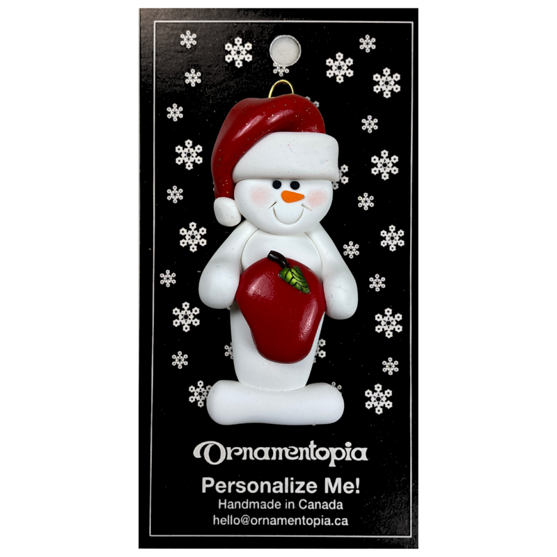 Ornament of white snowman in red hat holding a shiny red apple, on display backing card.