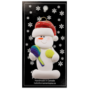 Ornament of white snowman in red hat holding a tennis paddle and ball, on display backing card.