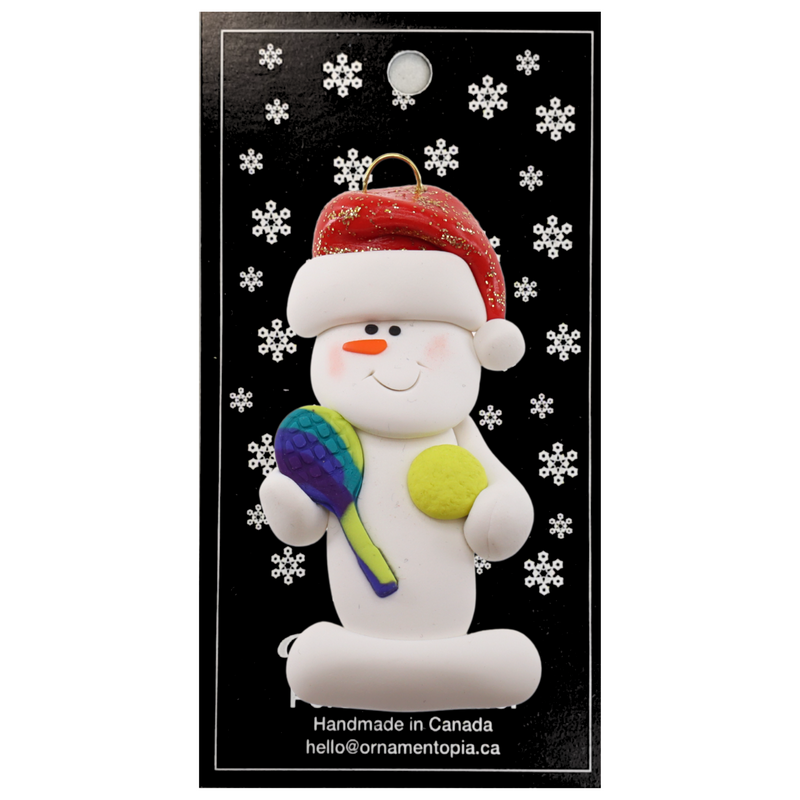 Ornament of white snowman in red hat holding a tennis paddle and ball, on display backing card.
