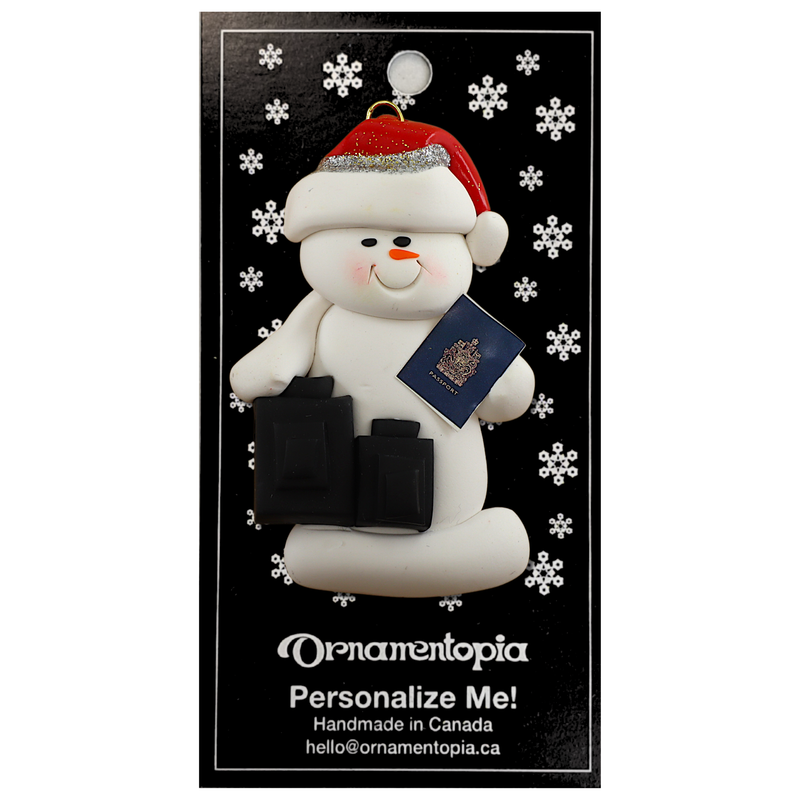 Ornament of white snowman in red hat holding a passport and suitcases, on display backing card.