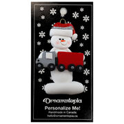 Ornament of white snowman in red hat holding a red semi truck, on display backing card.