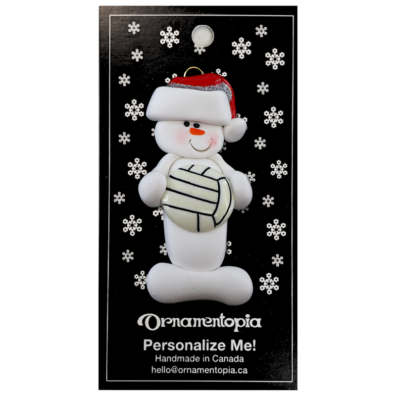 Ornament of white snowman in red hat holding a volleyball, on display backing card.