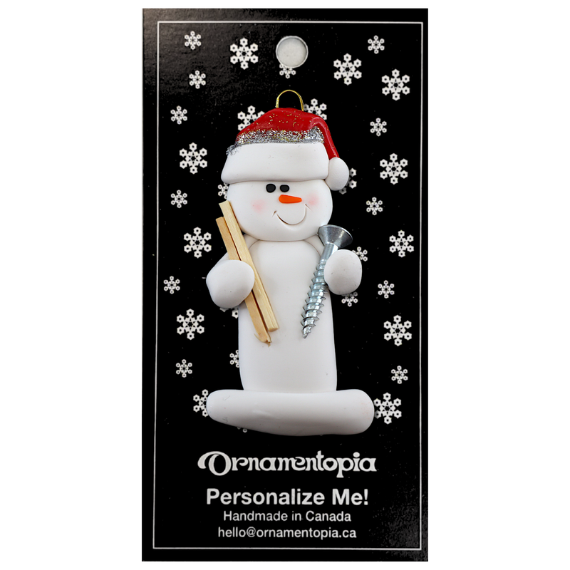 Ornament of white snowman in red hat holding a piece of wood and a screw, on display backing card.