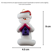 Ornament of white snowman in red hat holding a pink and purple birdhouse, graphed to show size (4.5x9cm).
