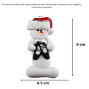 Ornament of white snowman in red hat holding black ballet shoes, graphed to show size (4.5x9cm).