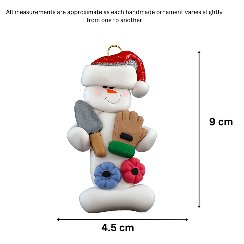 Ornament of white snowman in red hat holding a gardening glove and a shovel with flowers, graphed to show size (4.5x9cm).