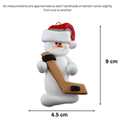 Ornament of white snowman in red hat holding a hockey stick and puck, graphed to show size (4.5x9cm).