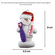 Ornament of white snowman in red hat holding a toothbrush and toothpaste, graphed to show size (4.5x9cm).