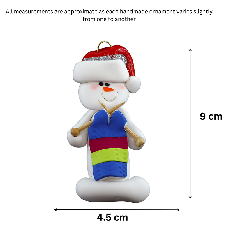 Ornament of white snowman in red hat holding an in-progress knitting project on knitting needles, graphed to show size (4.5x9cm).