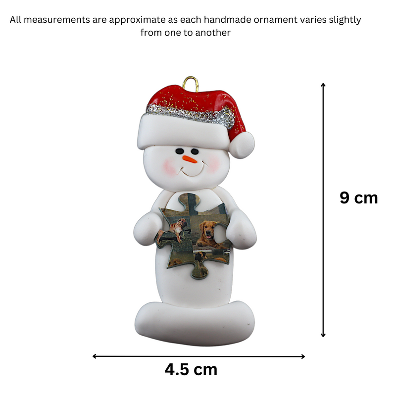 Ornament of white snowman in red hat holding a puzzle piece, graphed to show size (4.5x9cm).