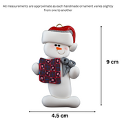 Ornament of white snowman in red hat holding a quilt block and pair of scissors, graphed to show size (4.5x9cm).