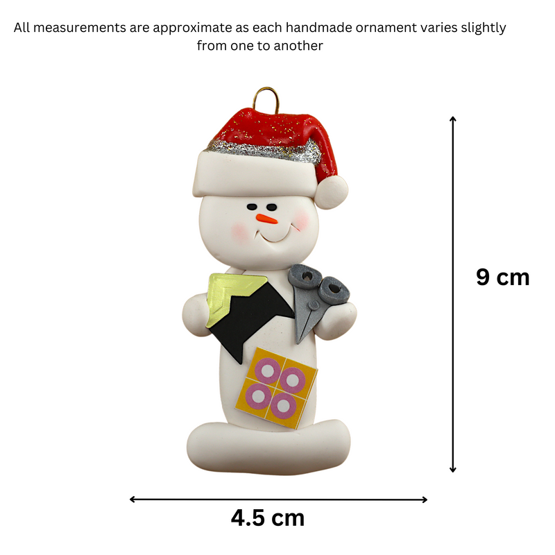 Ornament of white snowman in red hat holding a pair of scissors and various scrapbooking papers, graphed to show size (4.5x9cm).