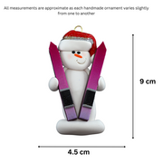 Ornament of white snowman in red hat holding a pair of vibrant skis, graphed to show size (4.5x9cm).