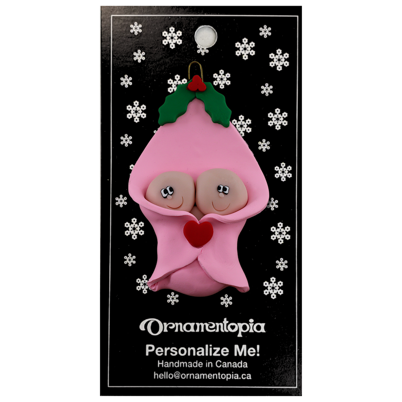 Ornament with twin infant heads poking out of a pink baby blanket, on display backing card.