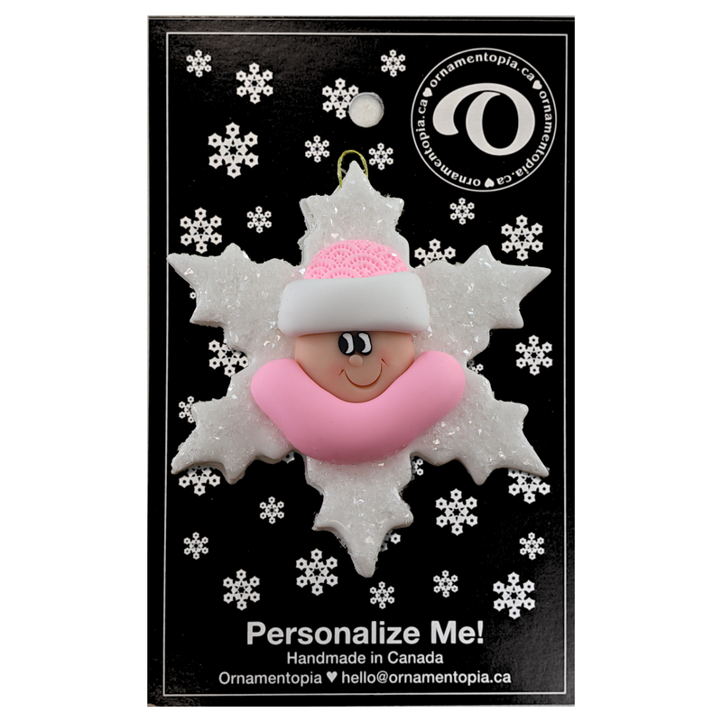 Baby ornament with infant face and pink hat on glittery white snowflake base, on display backing card.