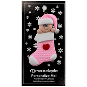 Ornament with infant head in a pink hat poking out of a pink stocking, on display backing card.