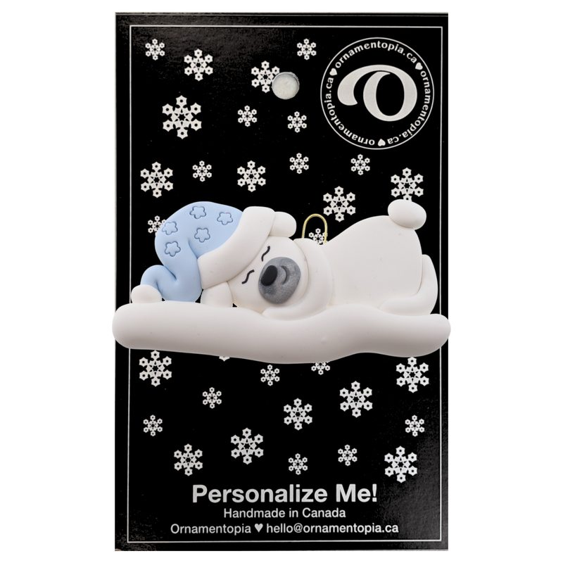 Ornament of white sleeping polar bear baby in a blue hat, on display backing card.