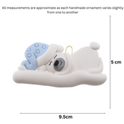 Ornament of white sleeping polar bear baby in a blue hat, graphed to show size (9.5x5cm).