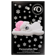 Ornament of white sleeping polar bear baby in a pink hat, on display backing card.