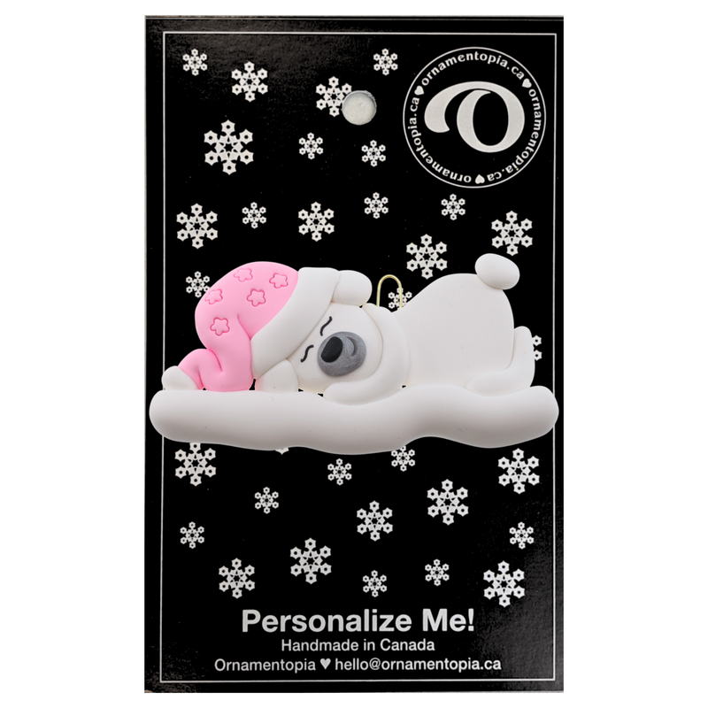 Ornament of white sleeping polar bear baby in a pink hat, on display backing card.