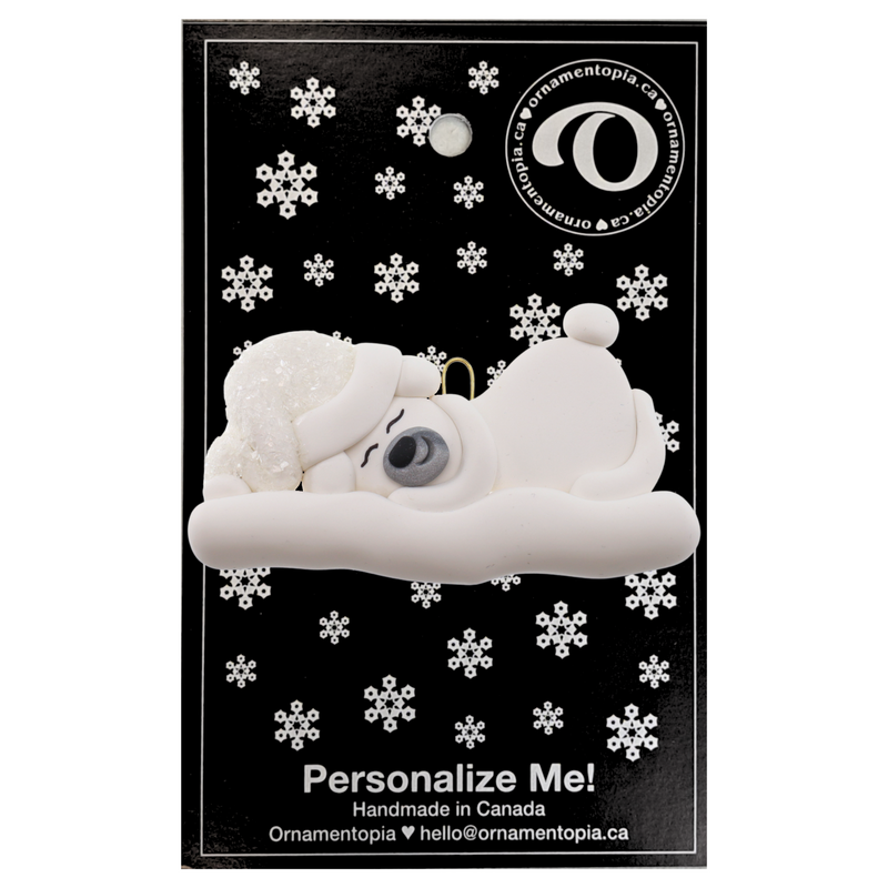 Ornament of white sleeping polar bear baby in a white sparkling hat, on display backing card.
