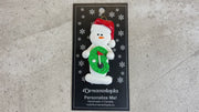 Ornament of white snowman in red hat holding a mini grass green with a golf ball and a flagstick, shown on backing card and Christmas tree.