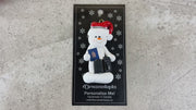 Ornament of white snowman in red hat holding a passport and suitcases, shown on backing card and Christmas tree.