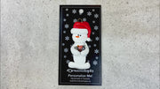 Ornament of white snowman in red hat holding several small rocks, shown on backing card and Christmas tree.