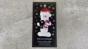 Ornament of white snowman in red hat holding a hairdryer and bottle of hair product, shown on backing card and Christmas tree.