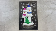 Family ornament with 2 white snowmen in bright scarves, with green Christmas tree, shown on backing card and Christmas tree.