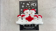 Family ornament with 6 white snowmen in red hats, gathered around a red maple leaf, shown on backing card and Christmas tree.
