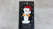 Ornament of white snowman in red hat holding orange cheerleader pom-poms, shown on backing card and Christmas tree.