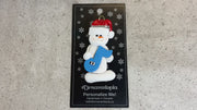 Ornament of white snowman in red hat holding a blue music note, shown on backing card and Christmas tree.