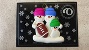 Family ornament with 3 white snowmen in bright hats with a football, shown on backing card and Christmas tree.