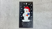 Ornament of white snowman in red hat holding cards and poker chips, shown on backing card and Christmas tree.