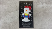 Ornament of white snowman in red hat holding a blue inflatable tube and yellow duck, shown on backing card and Christmas tree.