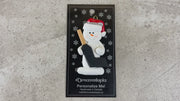 Ornament of white snowman in red hat holding a goalie stick, shown on backing card and Christmas tree.