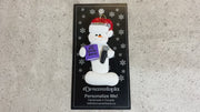 Ornament of white snowman in red hat holding a microphone and purple music book, shown on backing card and Christmas tree.