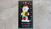 Ornament of white snowman in red hat holding various fruits, shown on backing card and Christmas tree.