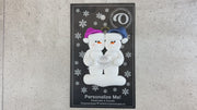 A couple’s ornament of two white snowmen holding a plastic diamond, shown on backing card and Christmas tree.