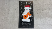 Ornament of white snowman in red hat holding an orange music note, shown on backing card and Christmas tree.
