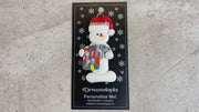 Ornament of white snowman in red hat holding a quilt block and pair of scissors, shown on backing card and Christmas tree.