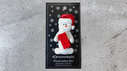 Ornament of white snowman in red hat holding a plastic building brick, shown on backing card and Christmas tree.